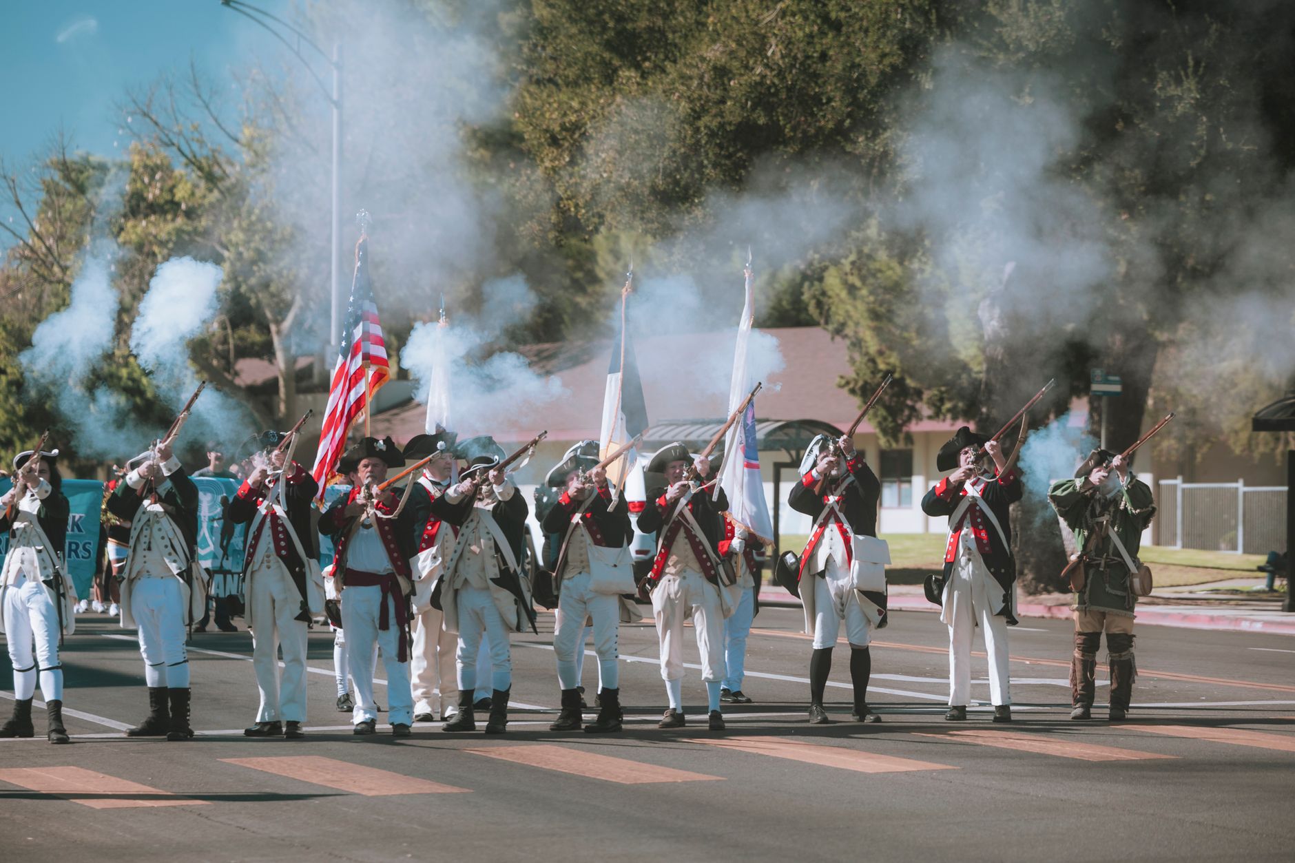 Participants in period costumes fire muskets in a dynamic historical reenactment parade.