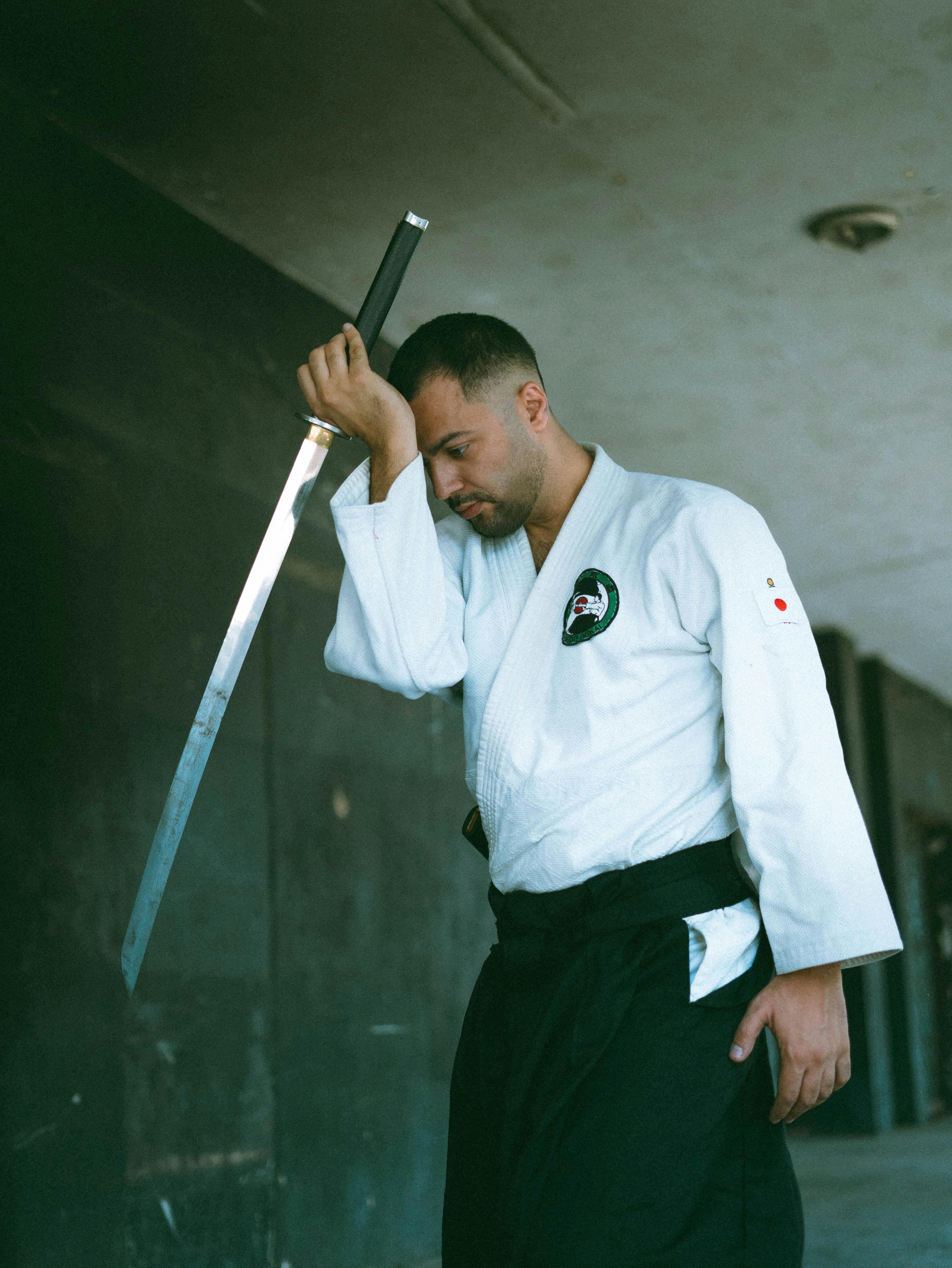 Martial artist in white uniform contemplatively holds a katana sword indoors.
