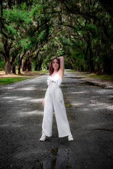 Woman in white jumpsuit posing in a scenic Savannah, Georgia road lined with moss-covered oak trees.