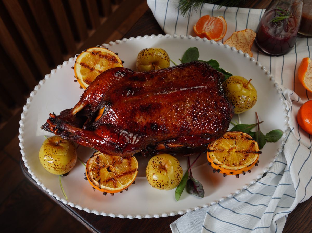 Whole roasted duck plated on a wooden board surrounded by grilled orange halves, grilled apples, thyme and rosemary — festive holiday presentation.