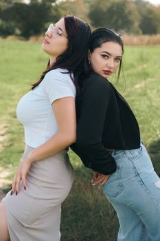 Two women posing back to back in a green field, wearing casual outfits.