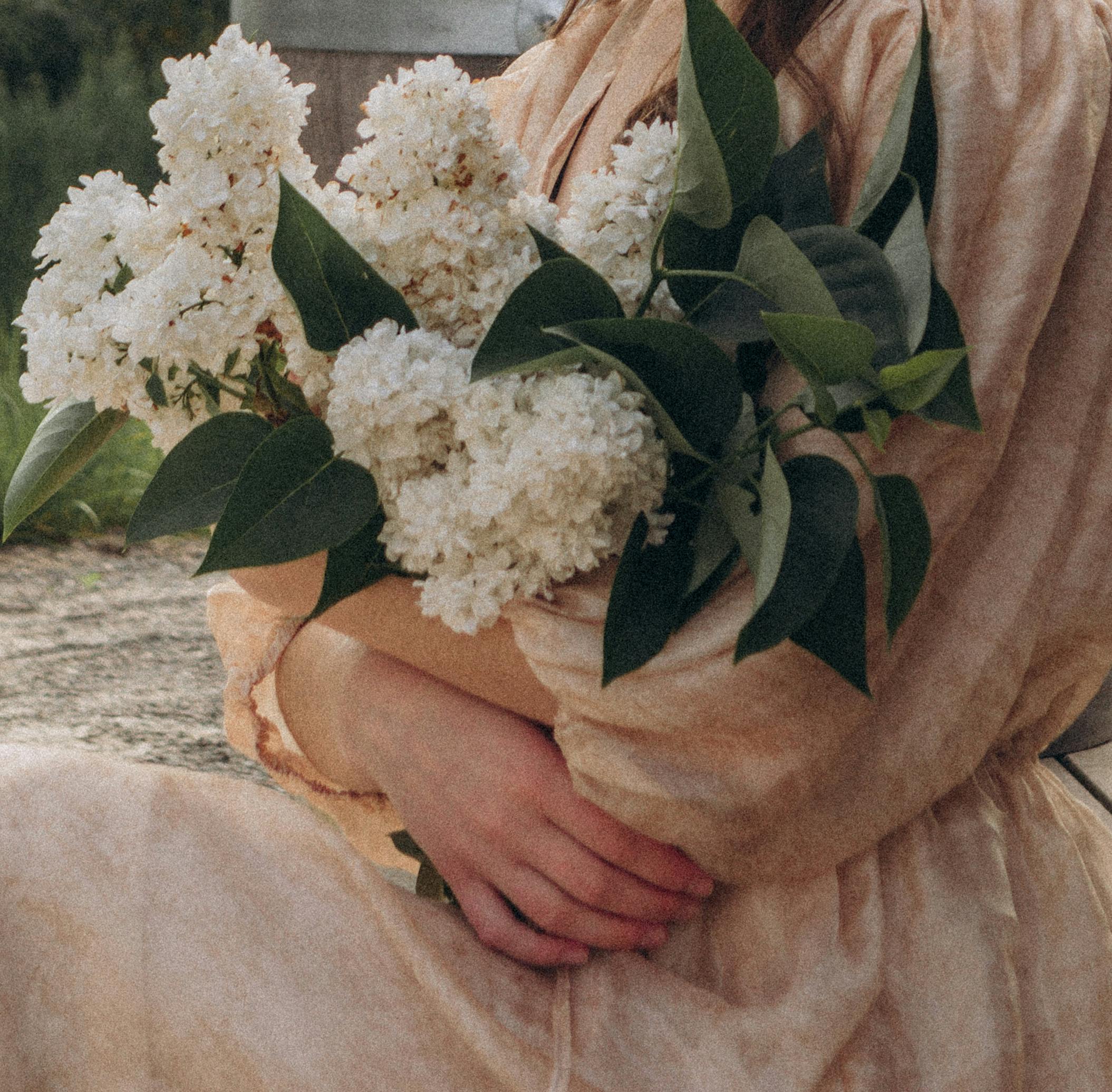 A woman in a beige dress holding a bouquet of white lilacs outdoors, capturing a serene moment.
