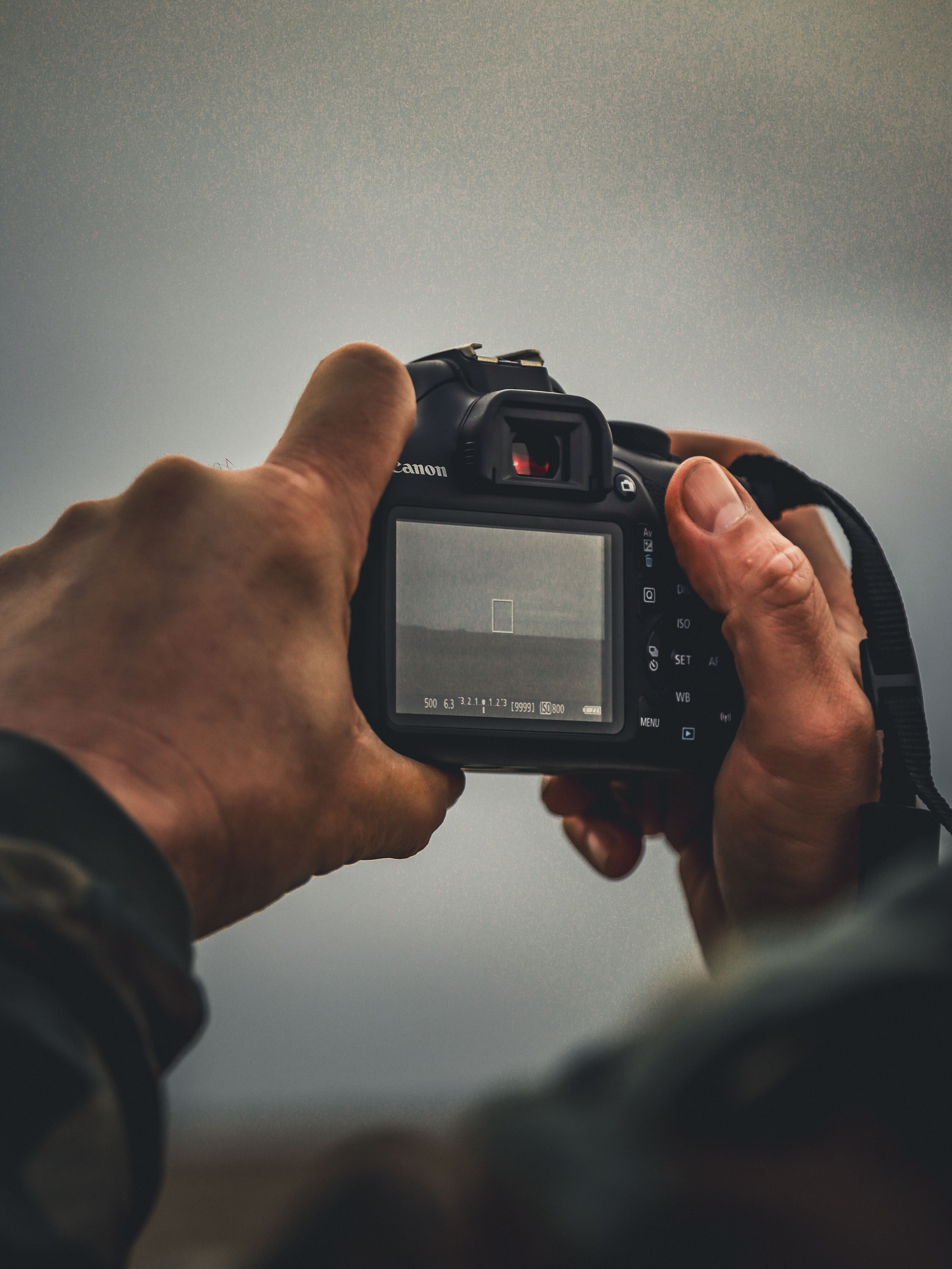 Hands holding a Canon camera, capturing an outdoor scene with a blurred background.