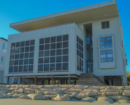 A modern coastal stilt house with large windows and sandbags for flood protection.