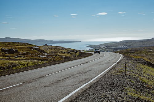 Empty asphalt road going through green valley of mountains with lonely car driving against blue sea harbor in sunlight