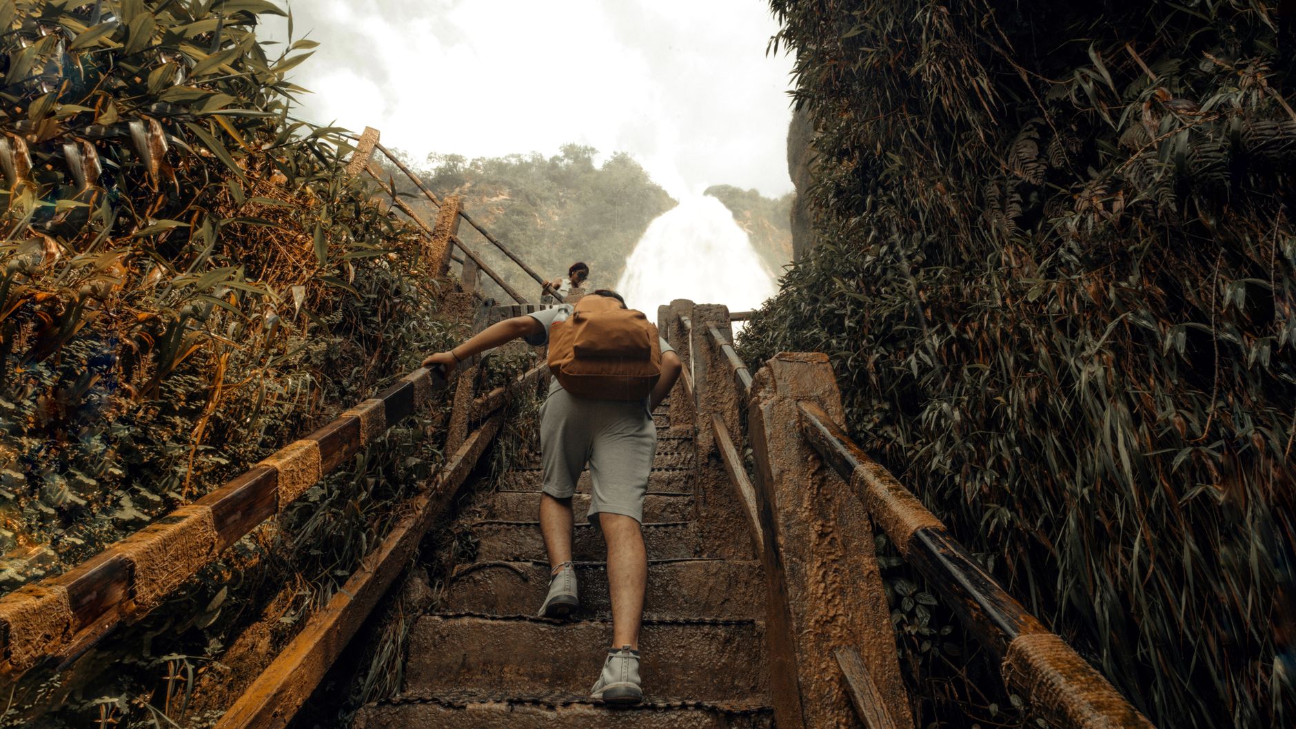 Man hikes up stone steps toward a large waterfall surrounded by lush greenery.