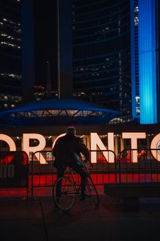 A cyclist pauses in front of the illuminated Toronto sign at City Hall, captured at night.