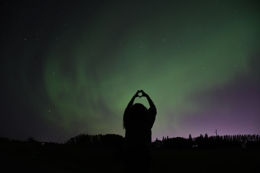 A person forms a heart with hands under the vibrant Northern Lights.