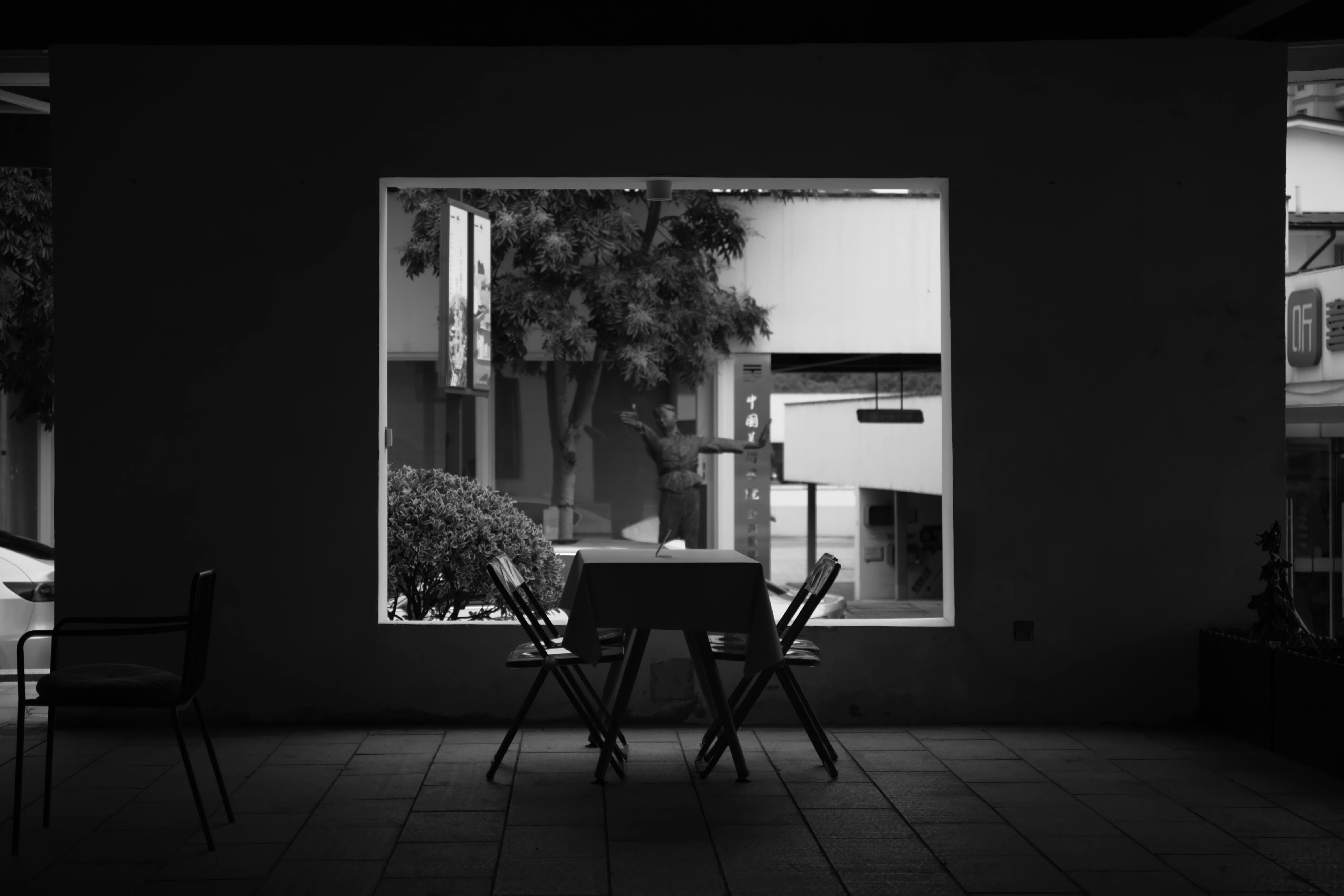 Kostenlos Ein Schwarzweißbild eines Straßencafés mit einem leeren Tisch und Stühlen, von drinnen betrachtet. Stock-Foto