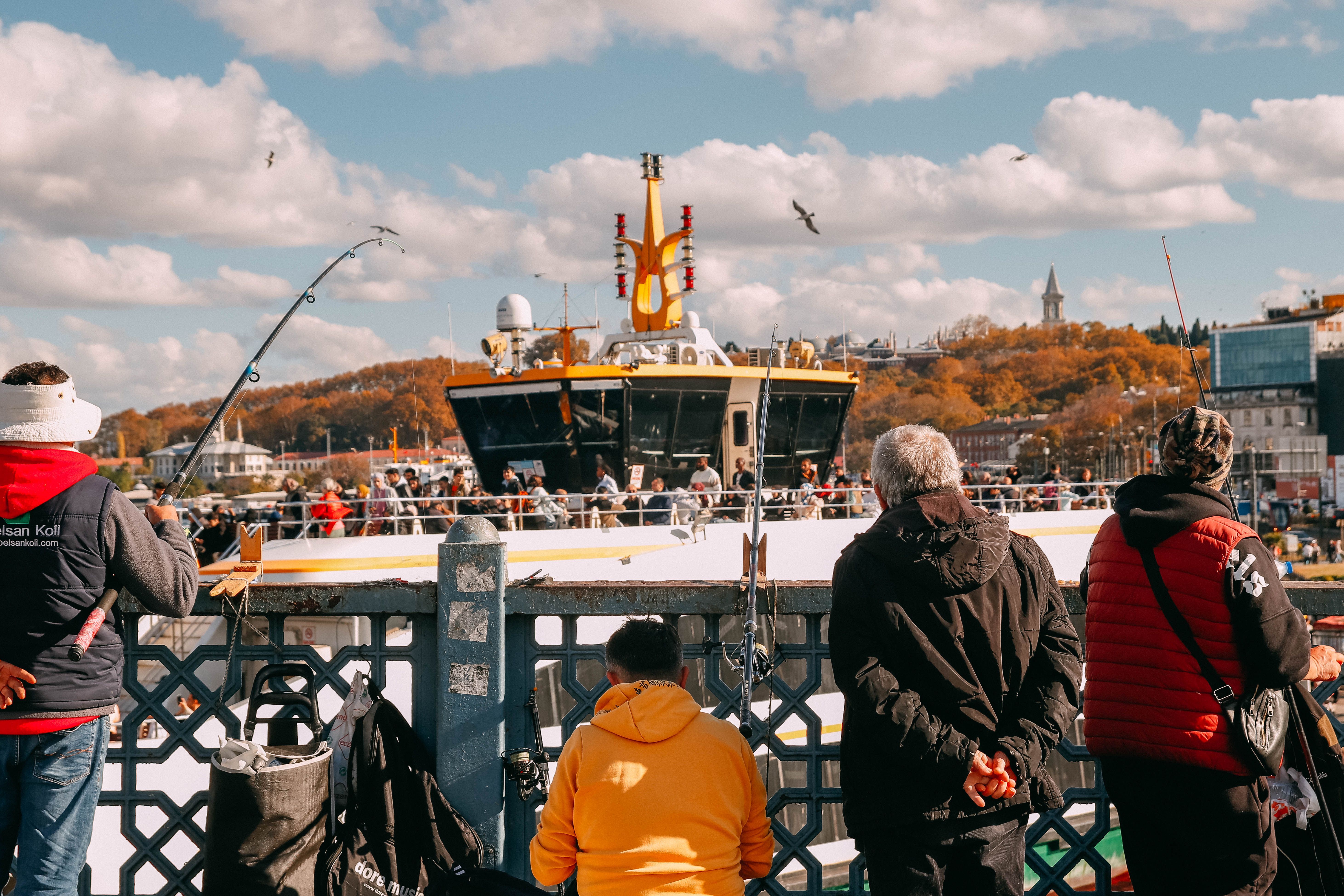 Fishermen casting lines on Istanbul's Galata Bridge, with a ferry and autumn backdrop.