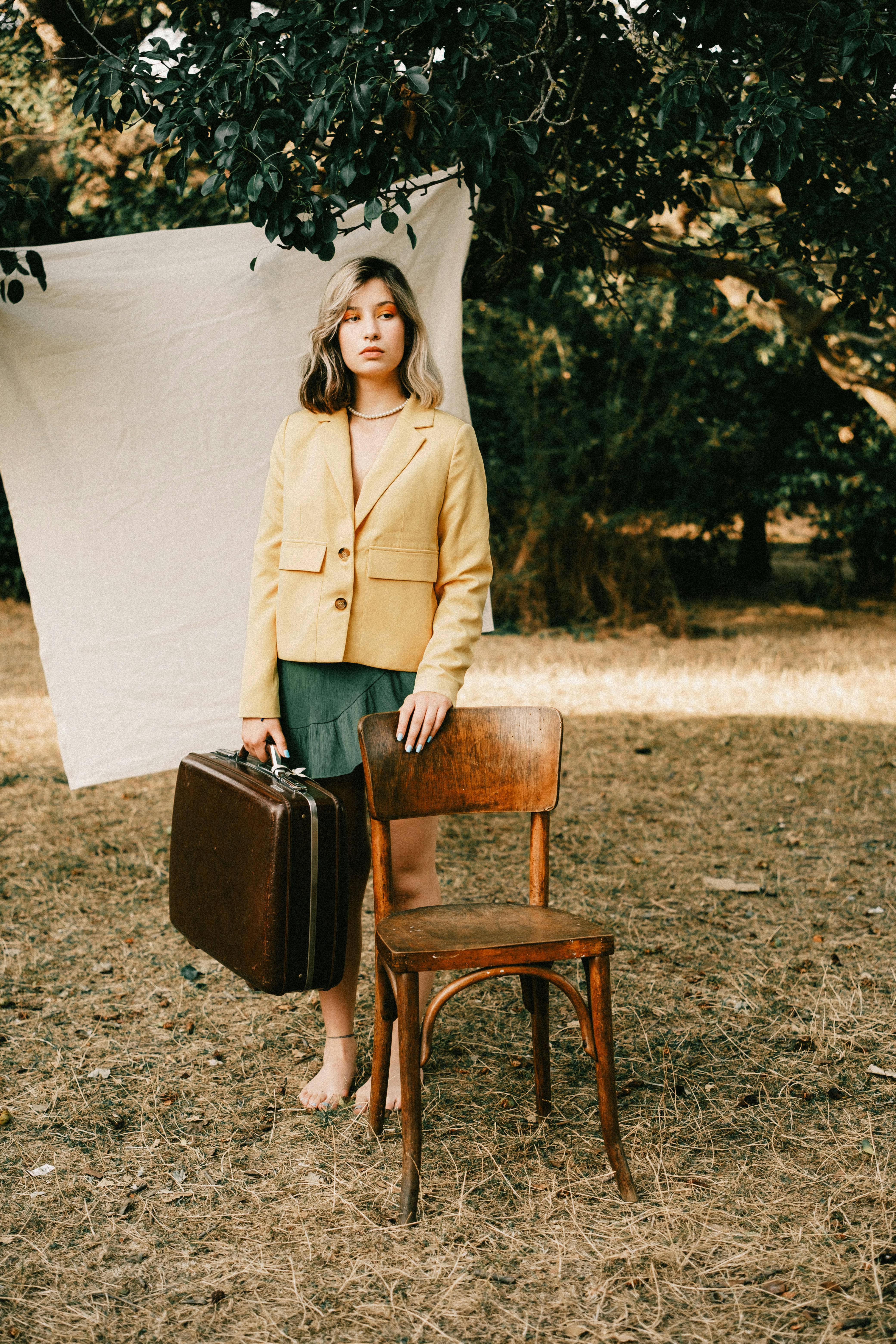 Young woman in nature holding a suitcase, standing by a chair.