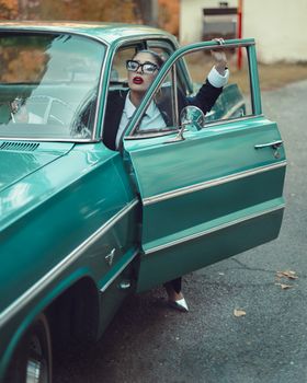 Fashionable woman in glasses gets out of a vintage turquoise car on a scenic autumn day.