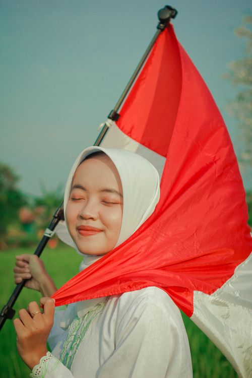 Free Young woman in hijab holding Indonesian flag, smiling outdoors. Stock Photo