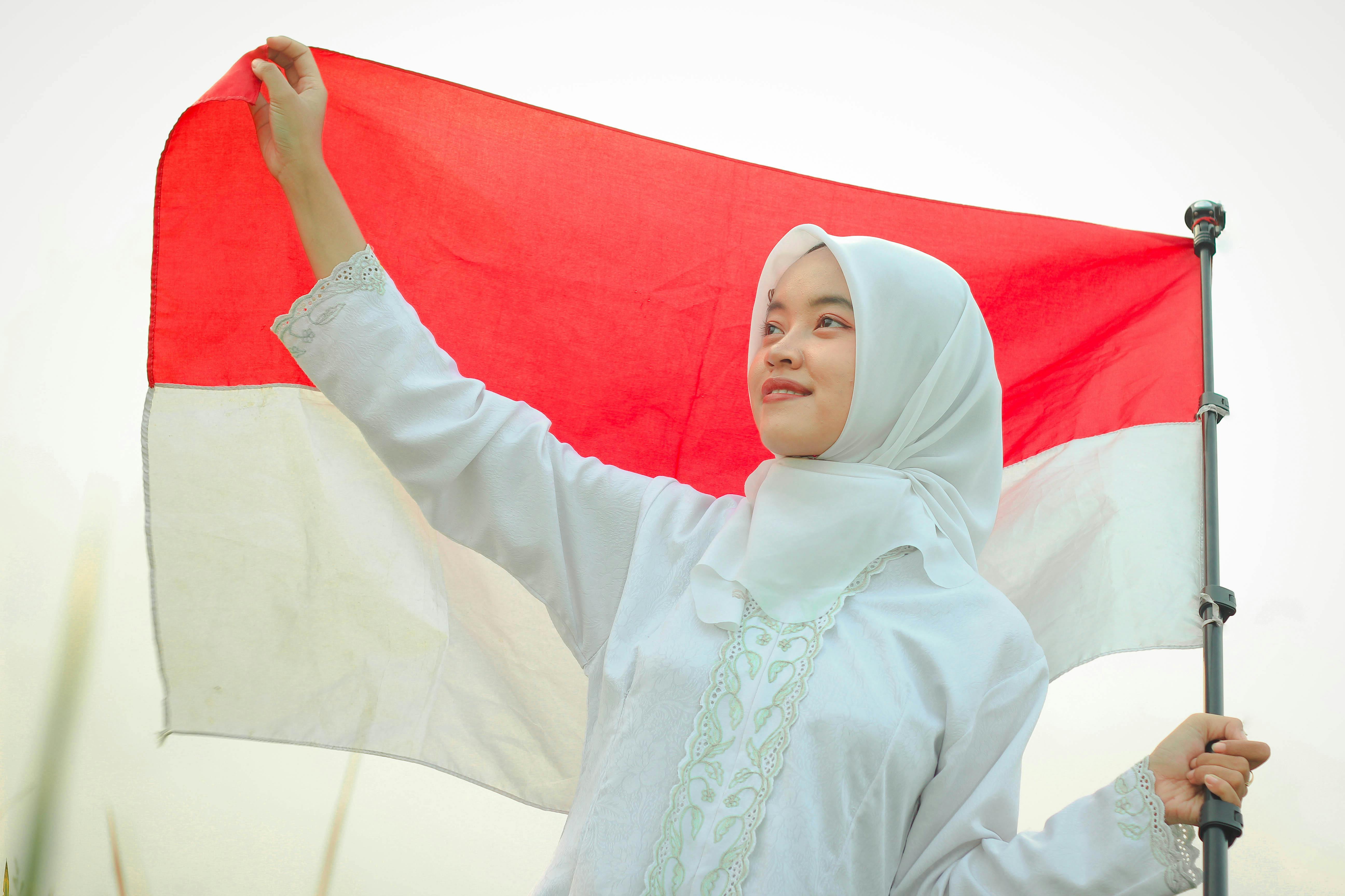 Portrait of a young woman in a hijab proudly holding the Indonesian flag outdoors during the day.