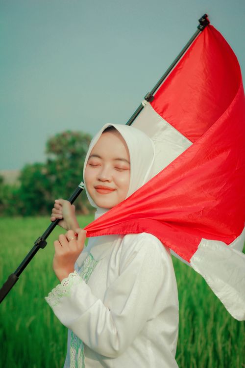 Free A young woman in a hijab joyously holding the Indonesian flag outdoors, symbolizing national pride. Stock Photo