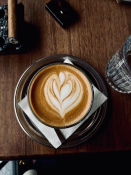 Latte with heart design on wooden table beside cigar.