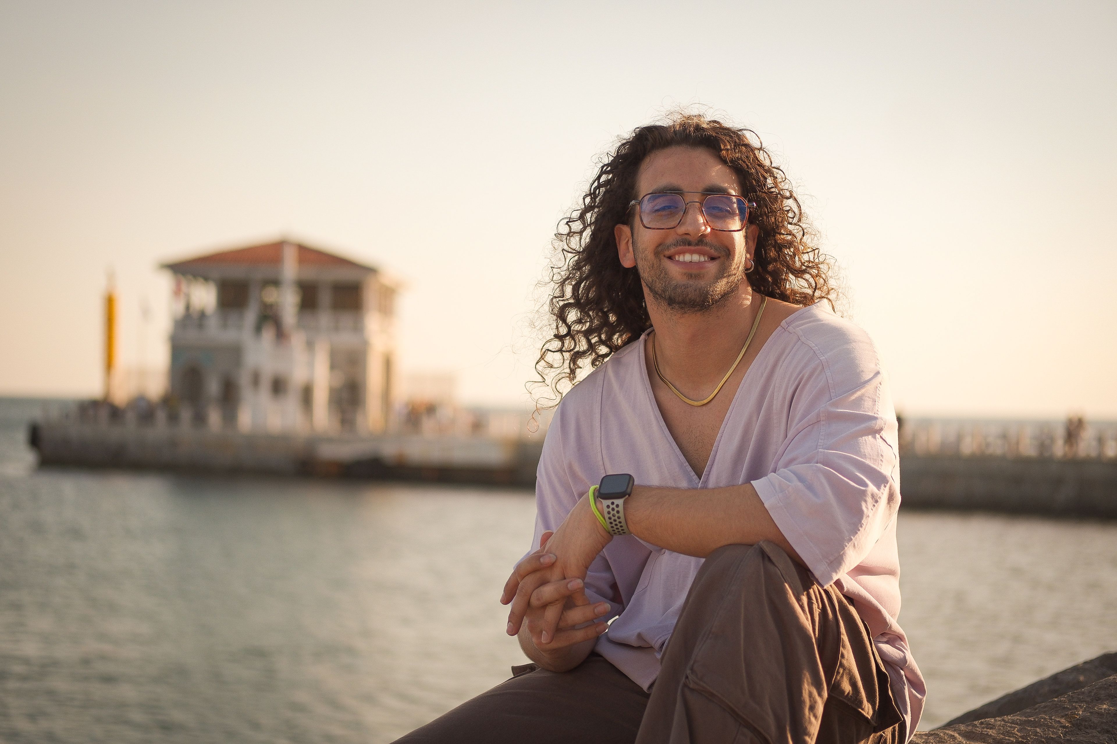 A cheerful man with curly hair enjoys the seaside in front of a historic building at sunset.
