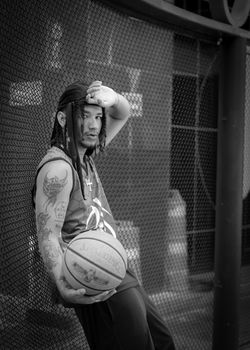 Black and white portrait of a stylish basketball player posing against a chain-link fence outdoors.