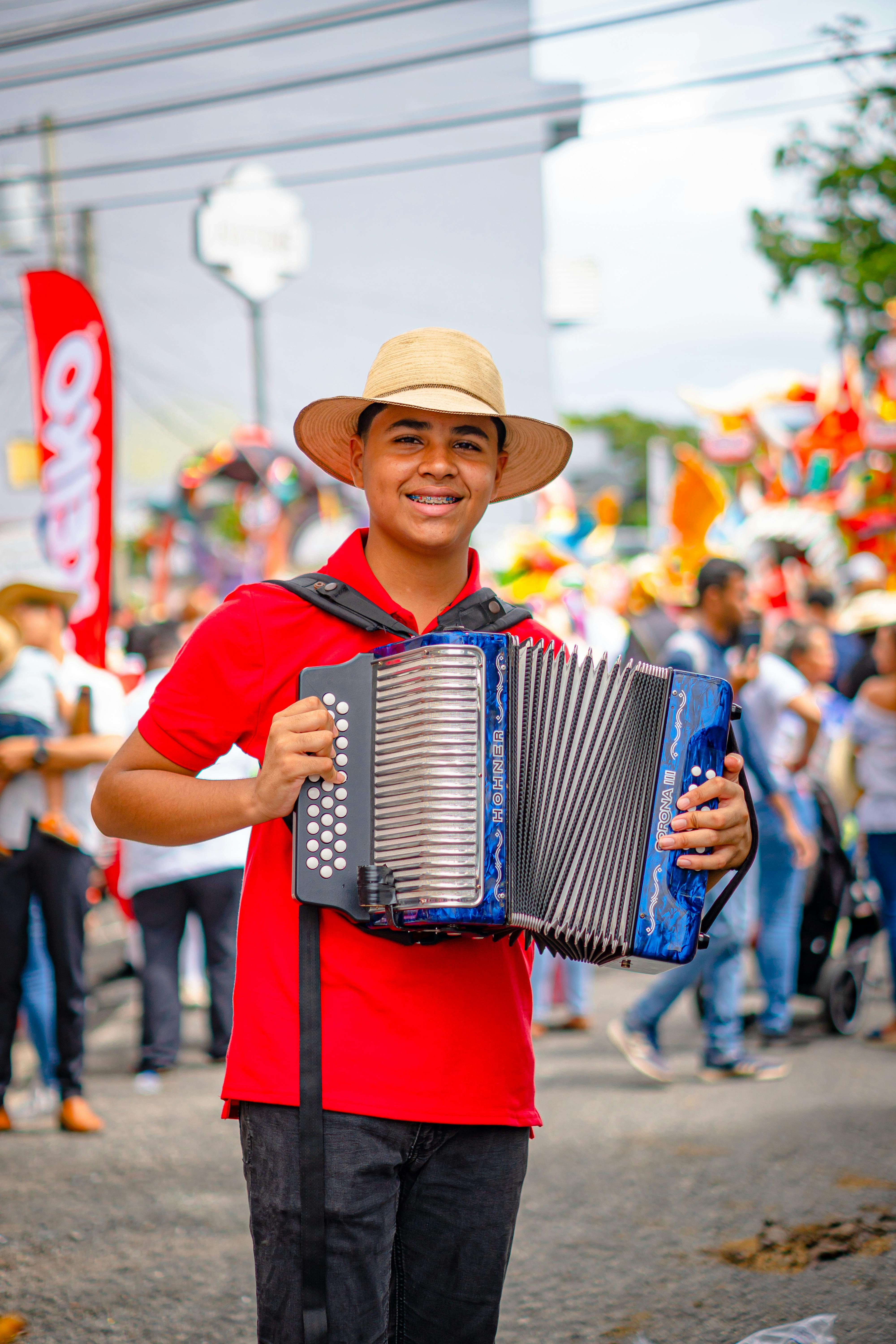 Smiling young man plays accordion at lively outdoor festival, wearing a traditional hat.