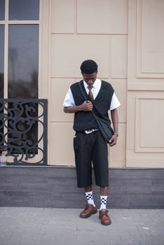 Young man in trendy urban outfit with checkered socks and a side bag standing against a beige wall.