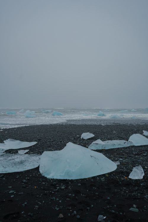 Free A serene icy beachfront with scattered icebergs on black pebbles under a misty sky. Stock Photo