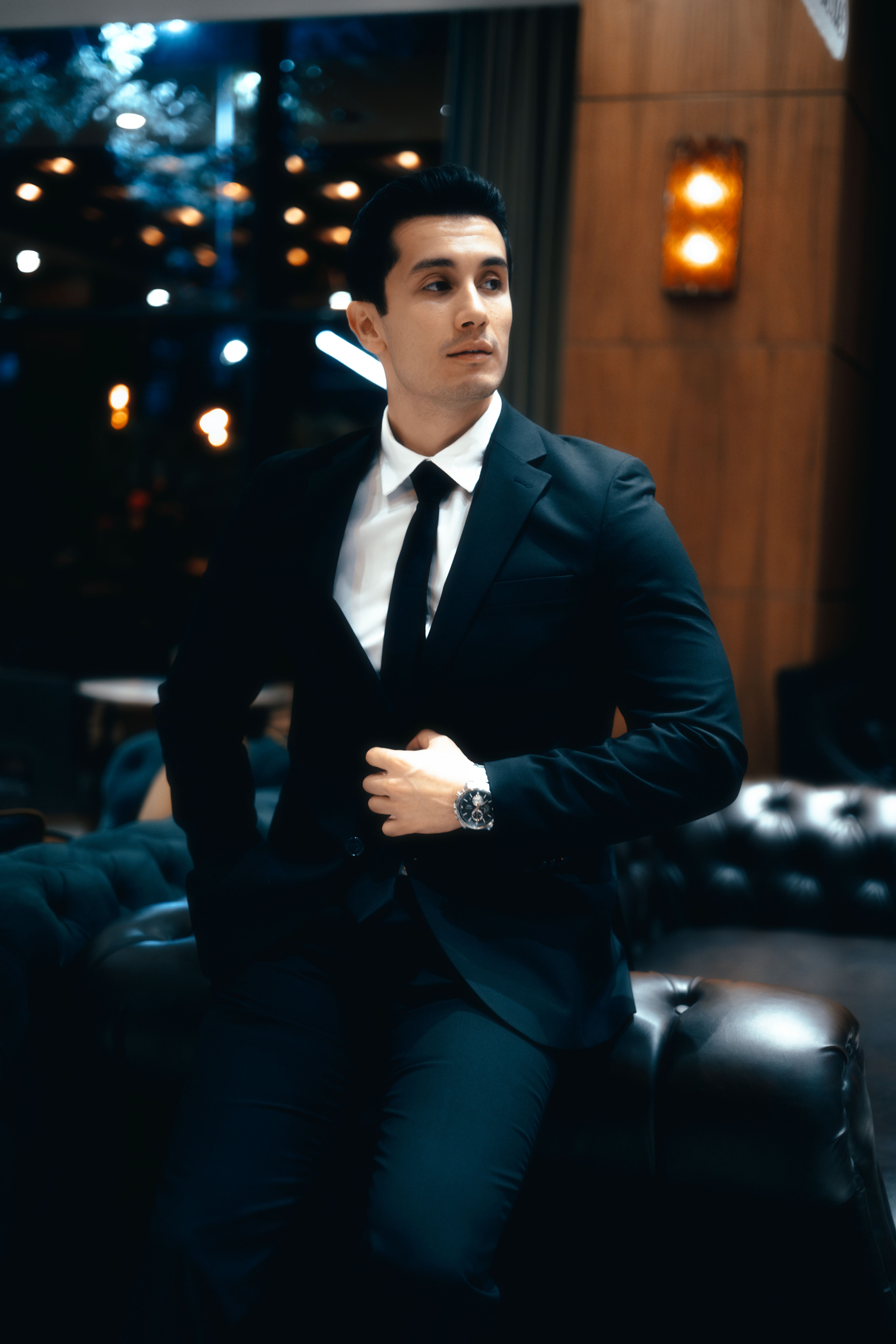A confident young man in a refined suit seated in an upscale lounge setting.