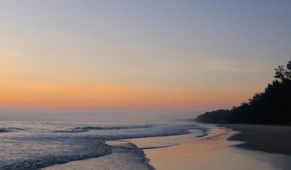 Tranquil Malaysian beach at sunrise with soft waves and an orange sky.