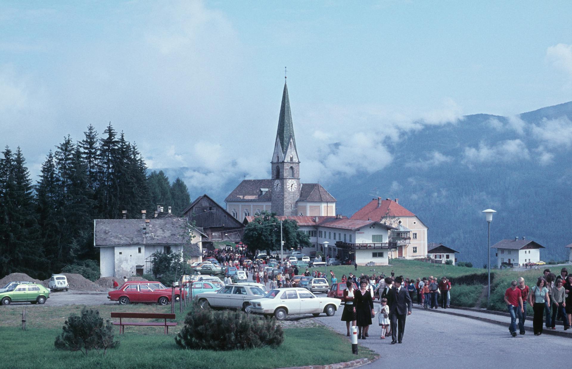 People gather near a picturesque church in Terento, Italy, surrounded by mountains.