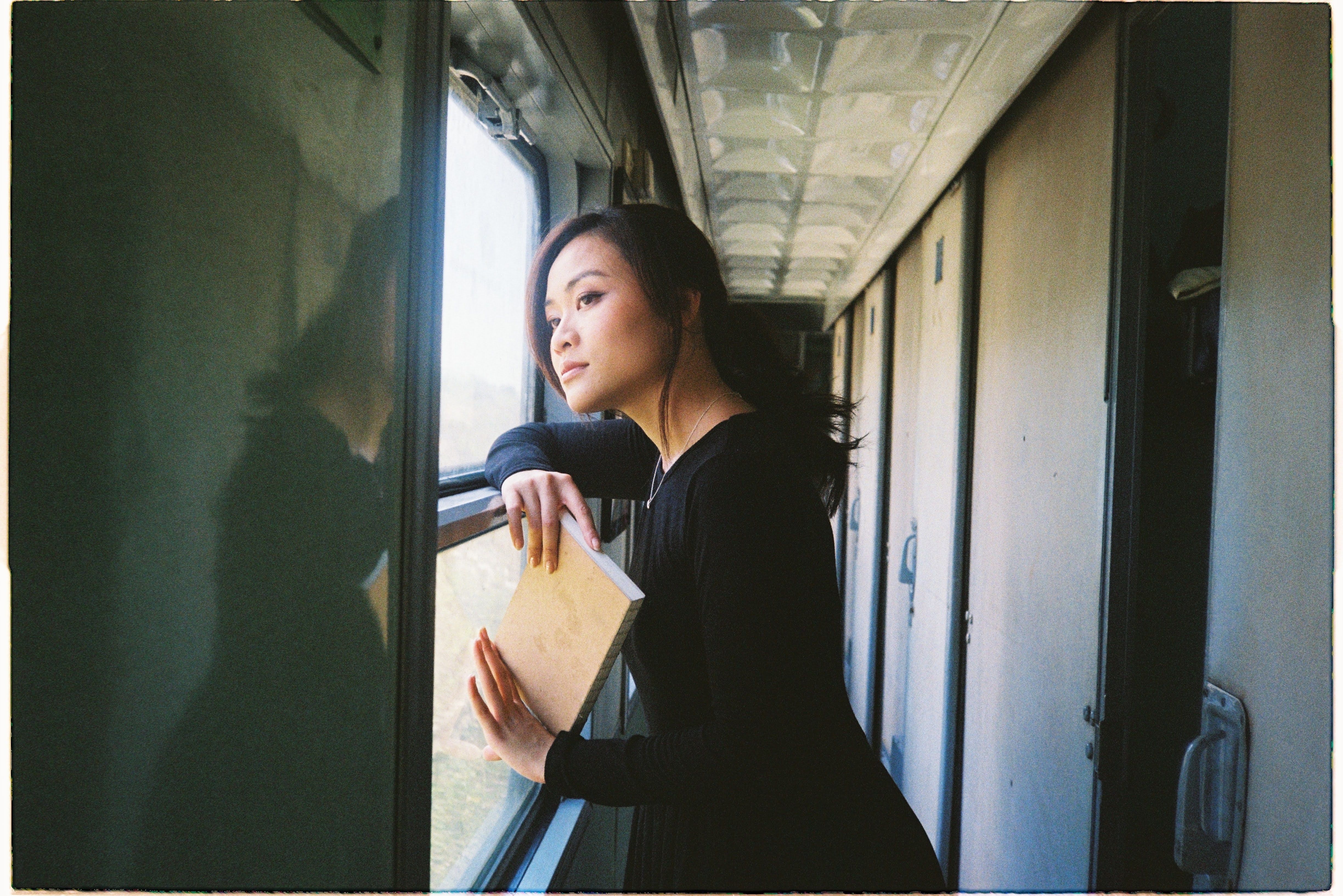 Woman gazing out train window, holding book in quiet reflection.