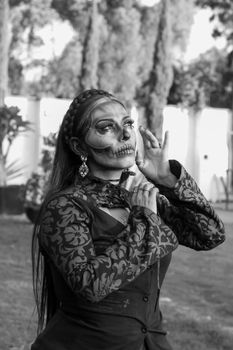 A woman with intricate Day of the Dead makeup poses elegantly outdoors in a black and white photograph.