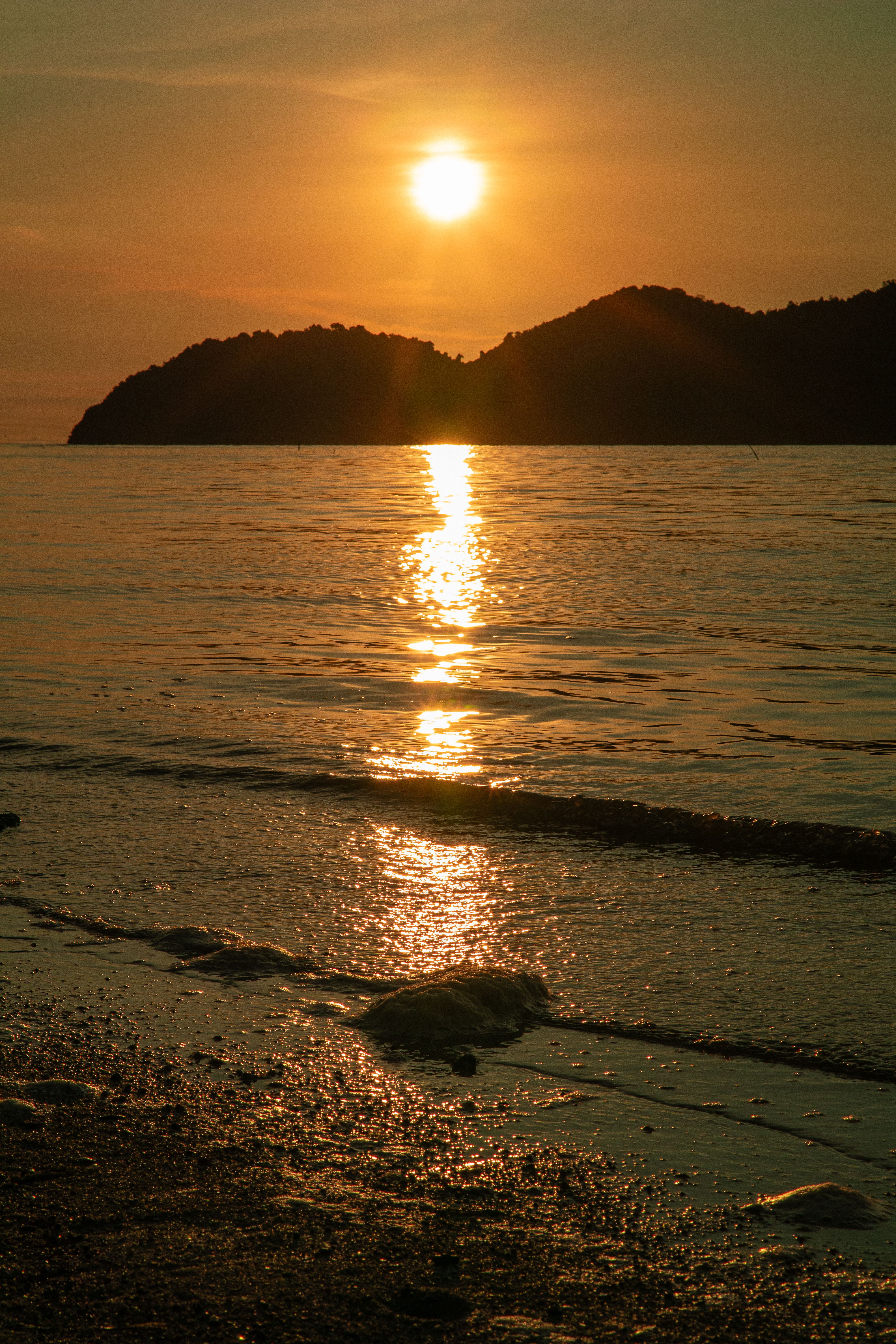 A tranquil sunset over Pulau Aman Beach, Penang, showcasing serene waters and silhouetted hills.