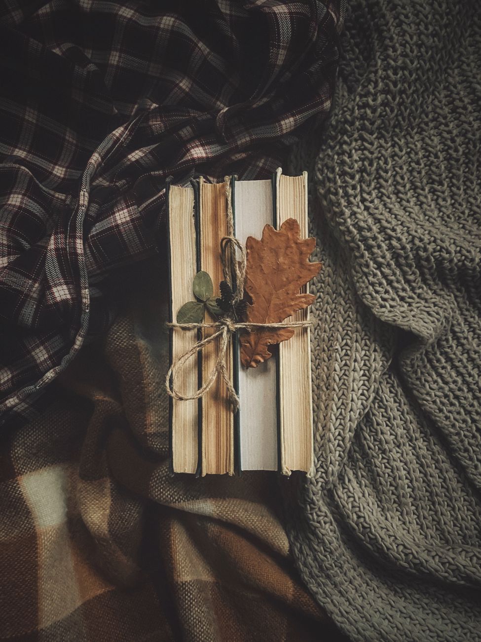 A stack of vintage books tied with twine, adorned with autumn leaves, surrounded by warm textiles.