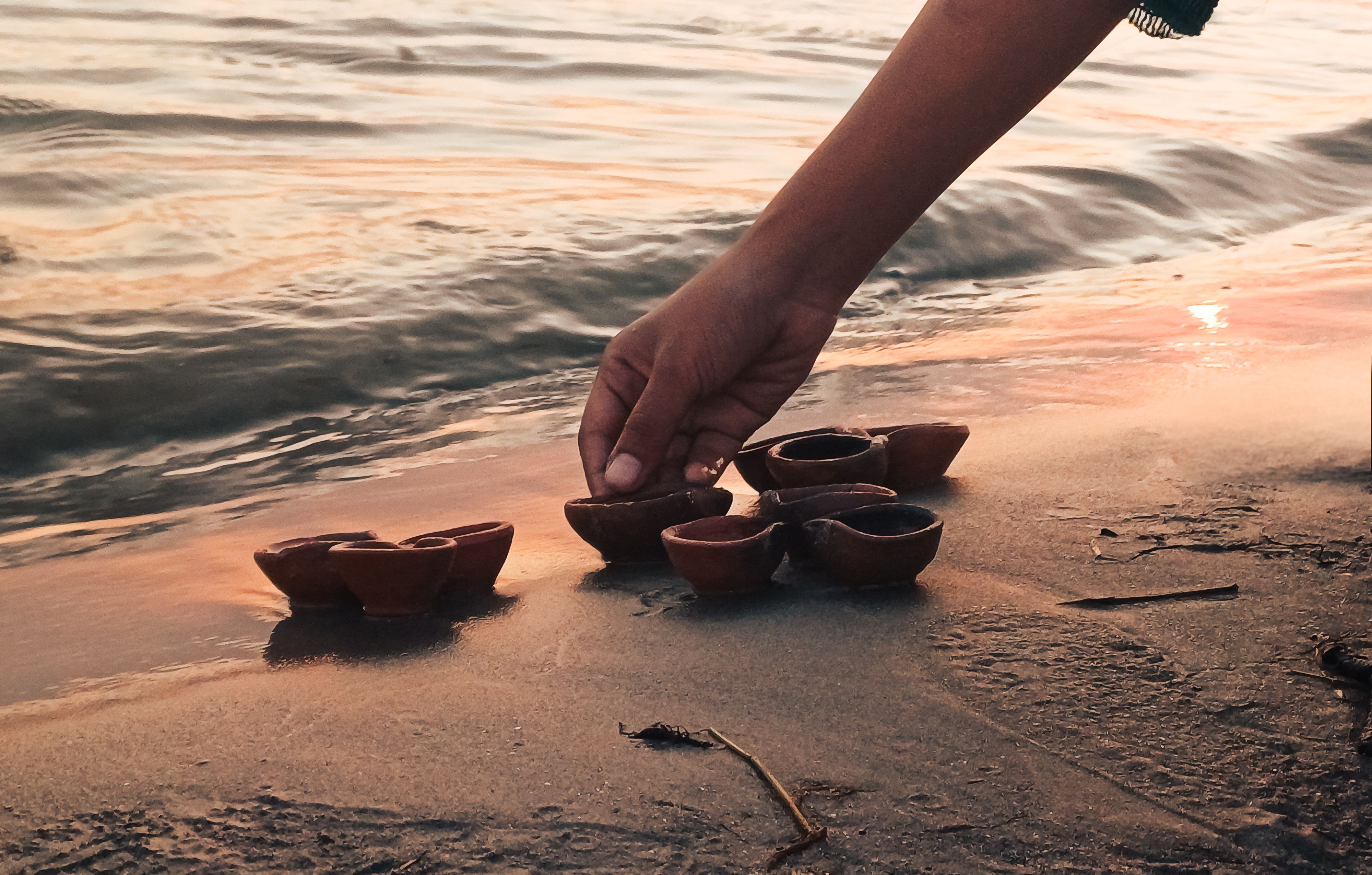 A hand arranging traditional diyas on a sandy riverside during sunset, capturing a serene moment. A hand arranging traditional diyas on a sandy riverside during sunset, capturing a serene moment.