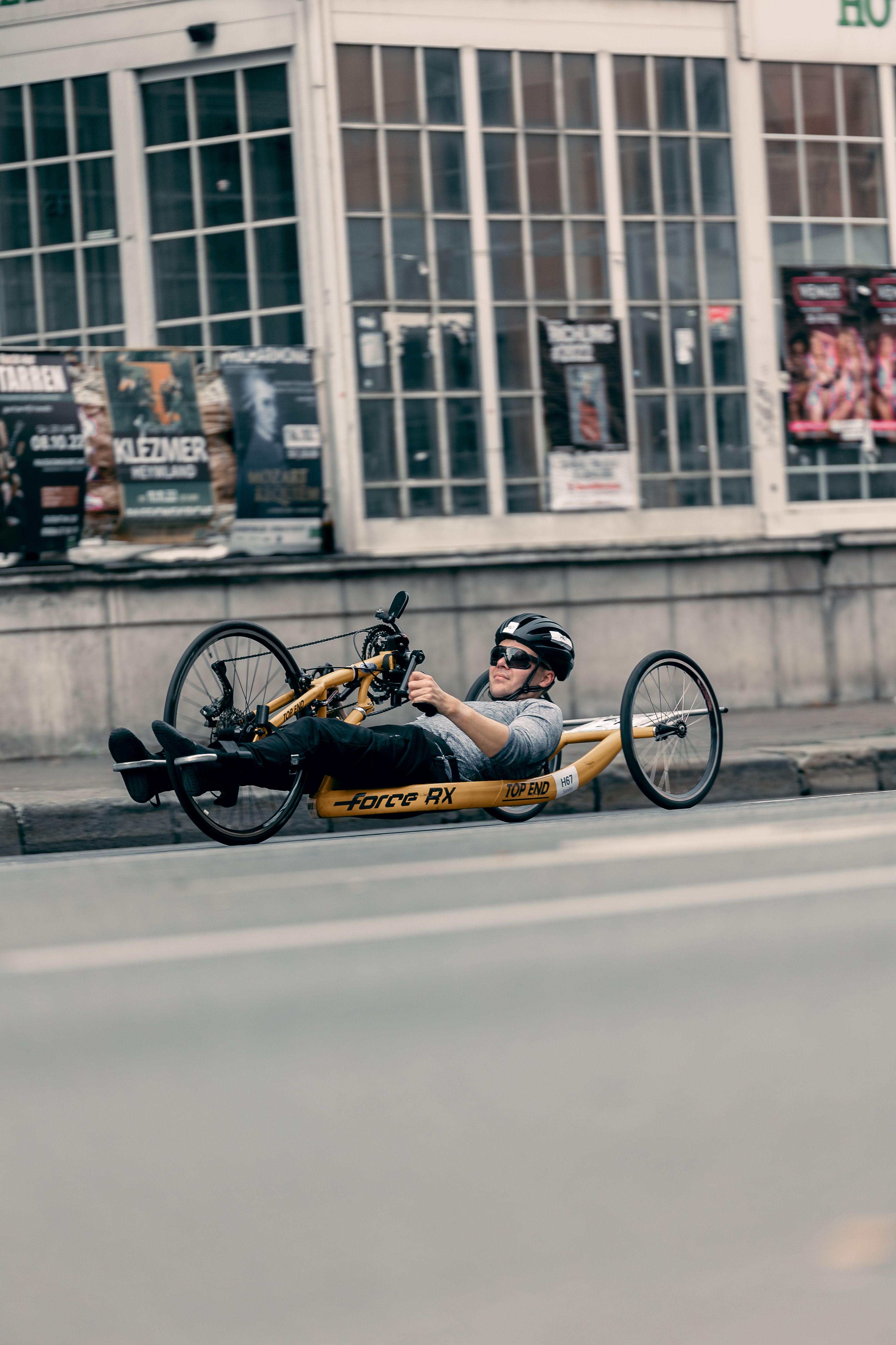 Adult man riding a handcycle along the street in urban setting, wearing helmet and sunglasses.