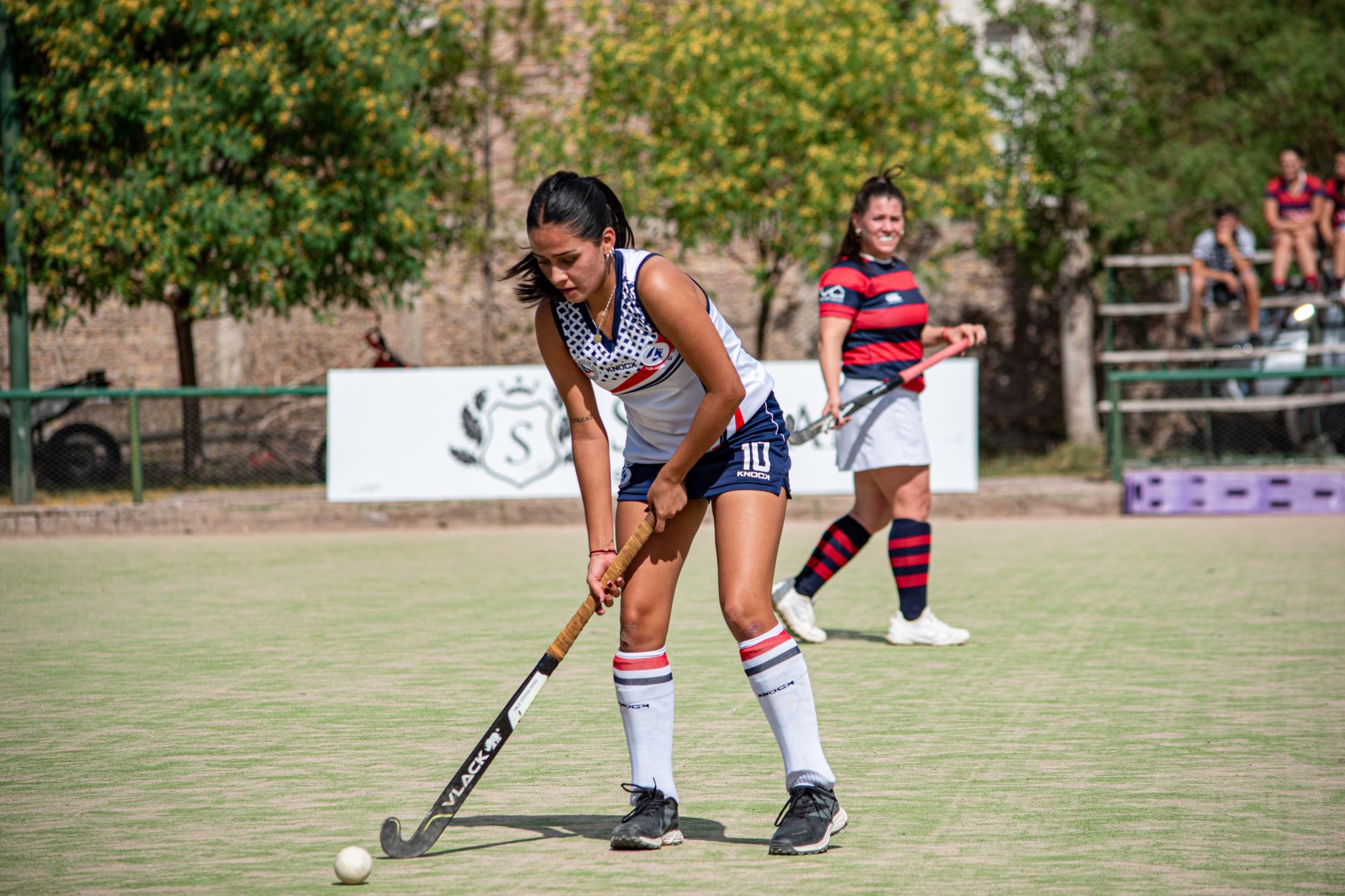 Female field hockey players competing during a sunny day match outdoors.