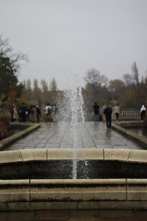 Free A tranquil fountain in a park with people strolling under a cloudy sky. Stock Photo