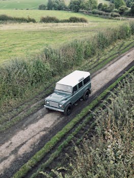 A vintage SUV travels on a dirt road through a lush countryside scene.