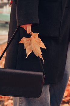 Close-up of a person holding a maple leaf and purse, symbolizing autumn fashion.