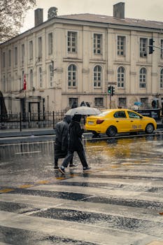 Two people with umbrellas cross a city street in the rain, yellow taxi in the background.