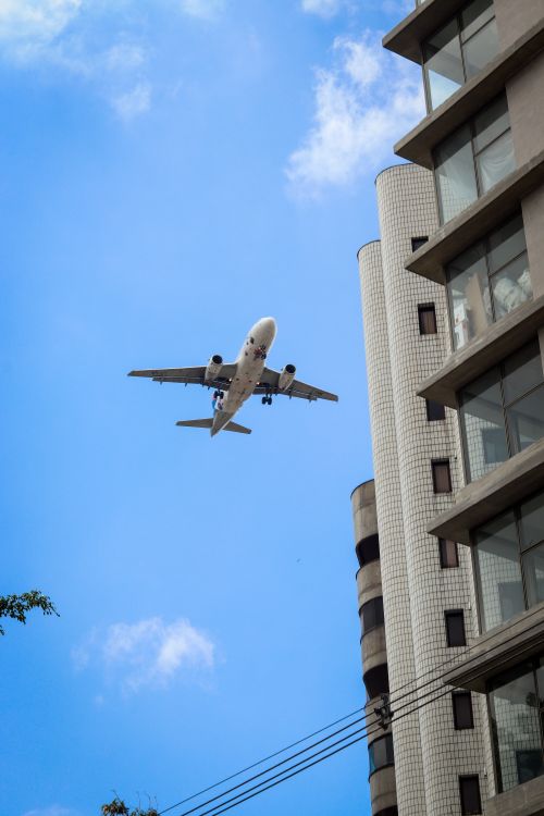 Free Commercial airplane soaring above city skyscraper against a clear blue sky. Stock Photo