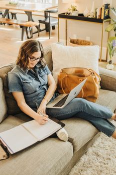 Woman in denim writing notes while working on a laptop with a leather tote bag beside her.