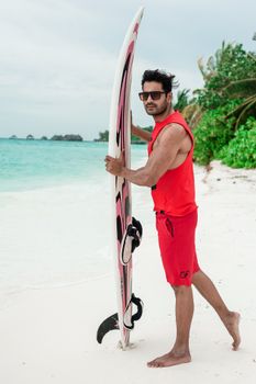 Man in red outfit holding a surfboard on a tropical Maldives beach, exuding beach style.