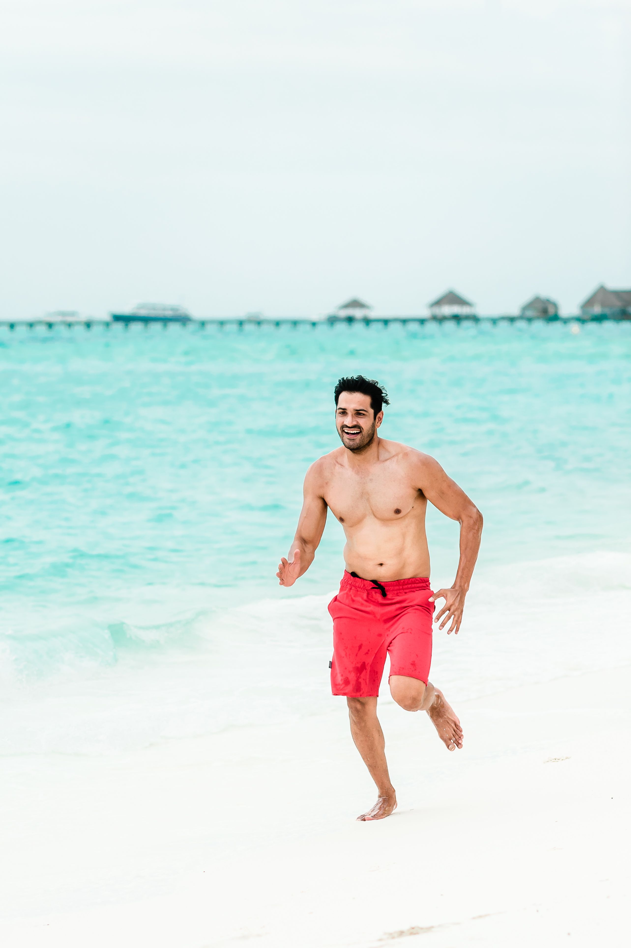 Smiling man in red shorts running on a Maldives beach with clear blue waters.
