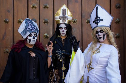Three adults in gothic cosplay with detailed skull makeup and bishop costumes in front of a wooden door.