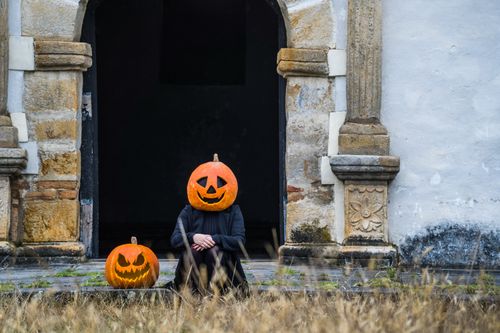 Free Person with pumpkin head sits by historic stone building, creating a Halloween ambiance. Stock Photo