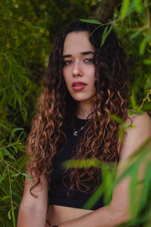 Free Portrait of a young woman with curly hair surrounded by green foliage. Stock Photo