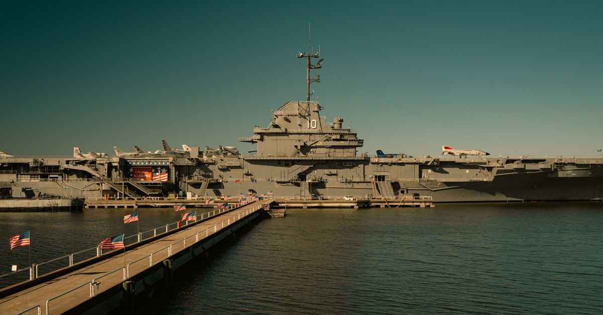 View of USS Yorktown aircraft carrier docked at harbor with walkway and flags.