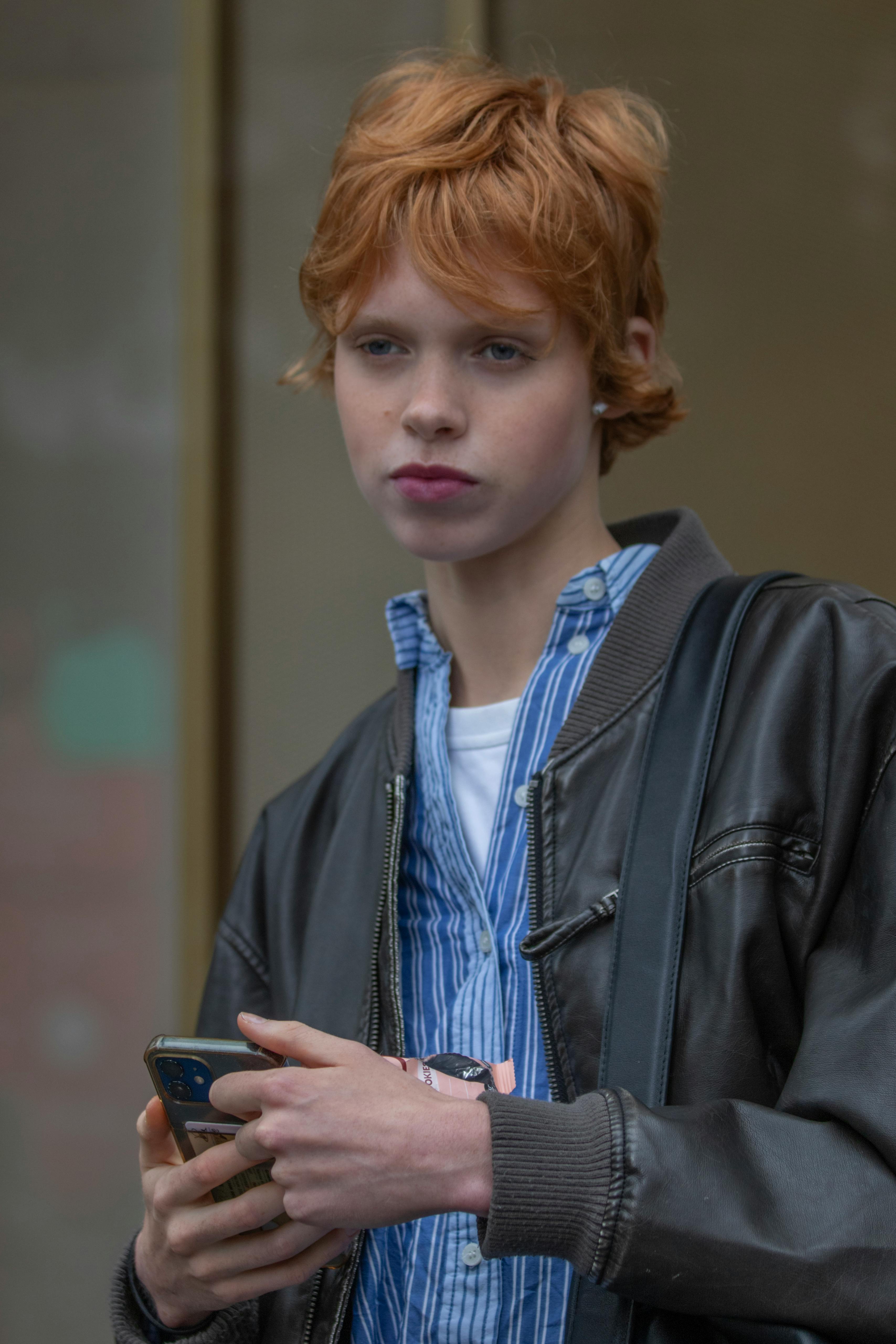 Kostenlos Teenager mit roten Haaren hält sein Handy in einer New Yorker Straße und trägt legere Kleidung. Stock-Foto