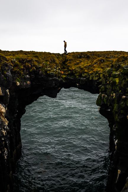 A solitary traveler walks on a rock arch over the ocean in Iceland, showcasing nature's raw beauty.