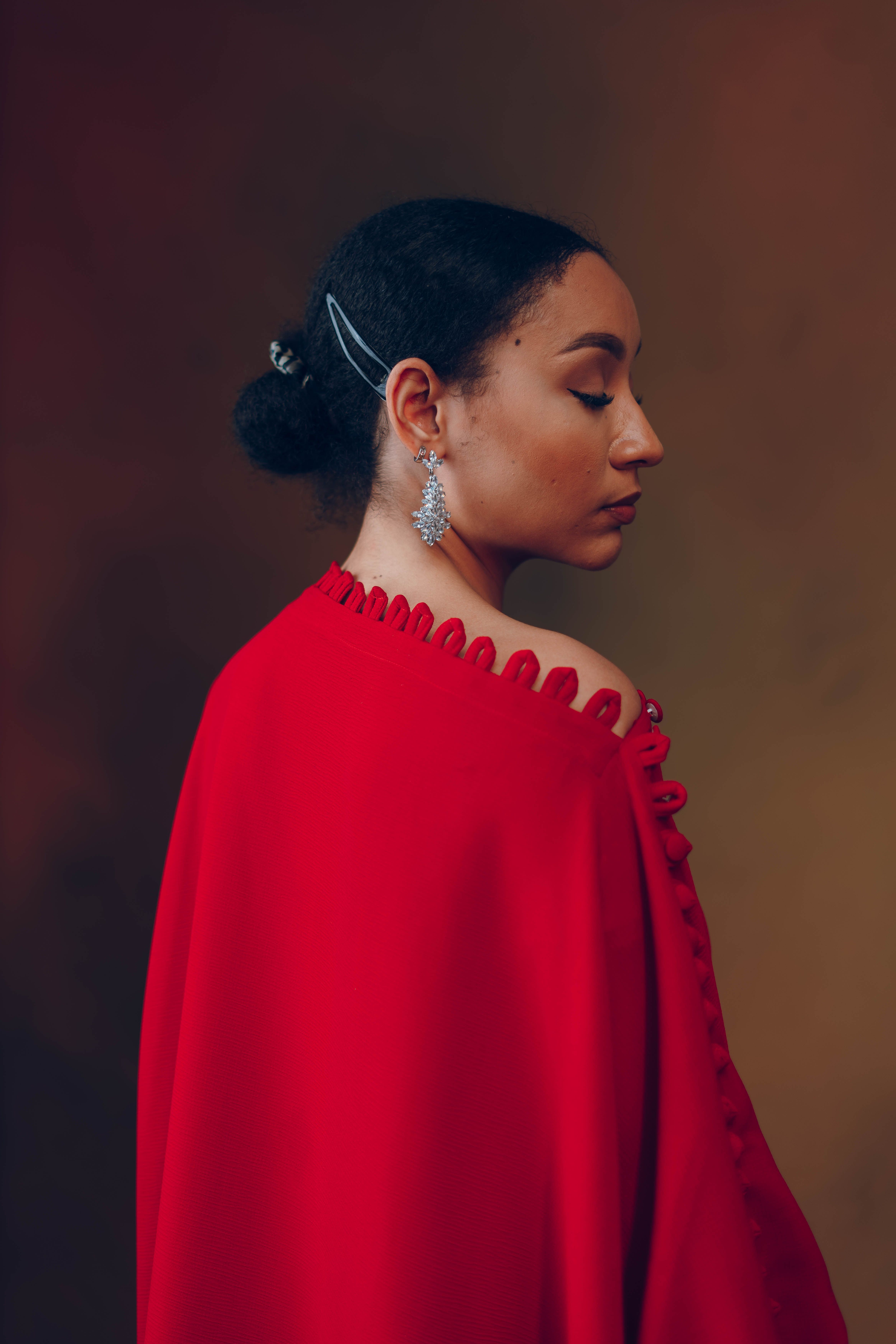 Side profile of a woman in a red cape with elegant earrings, on a muted background.